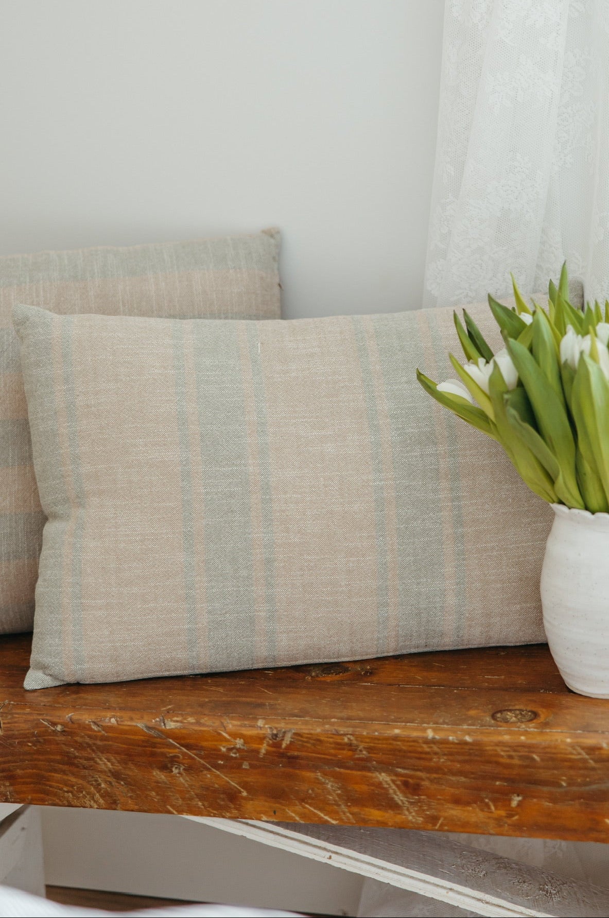 Vase with greenery on a wooden surface next to a striped pillow against a neutral wall.