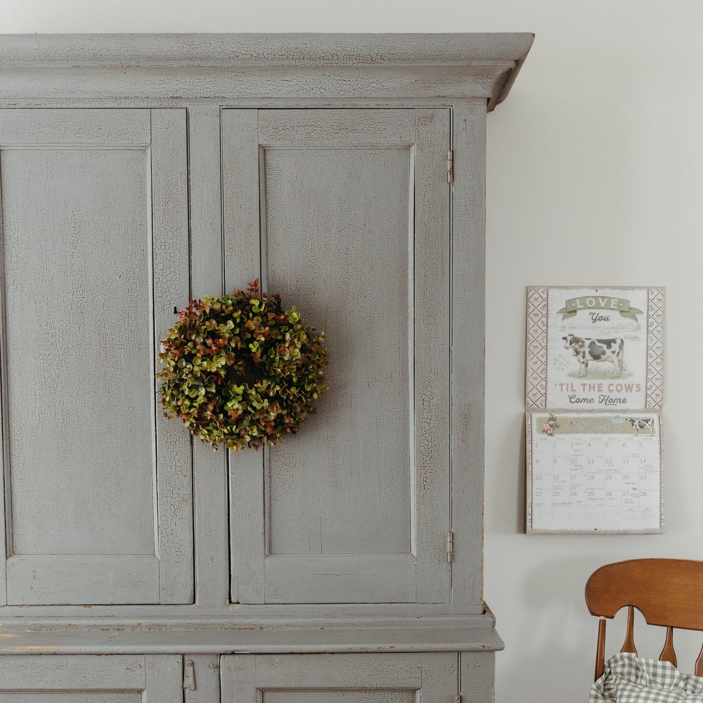 Gray wooden cabinet with a wreath on top, wooden chair with plaid cushion, and wall decor in a room.