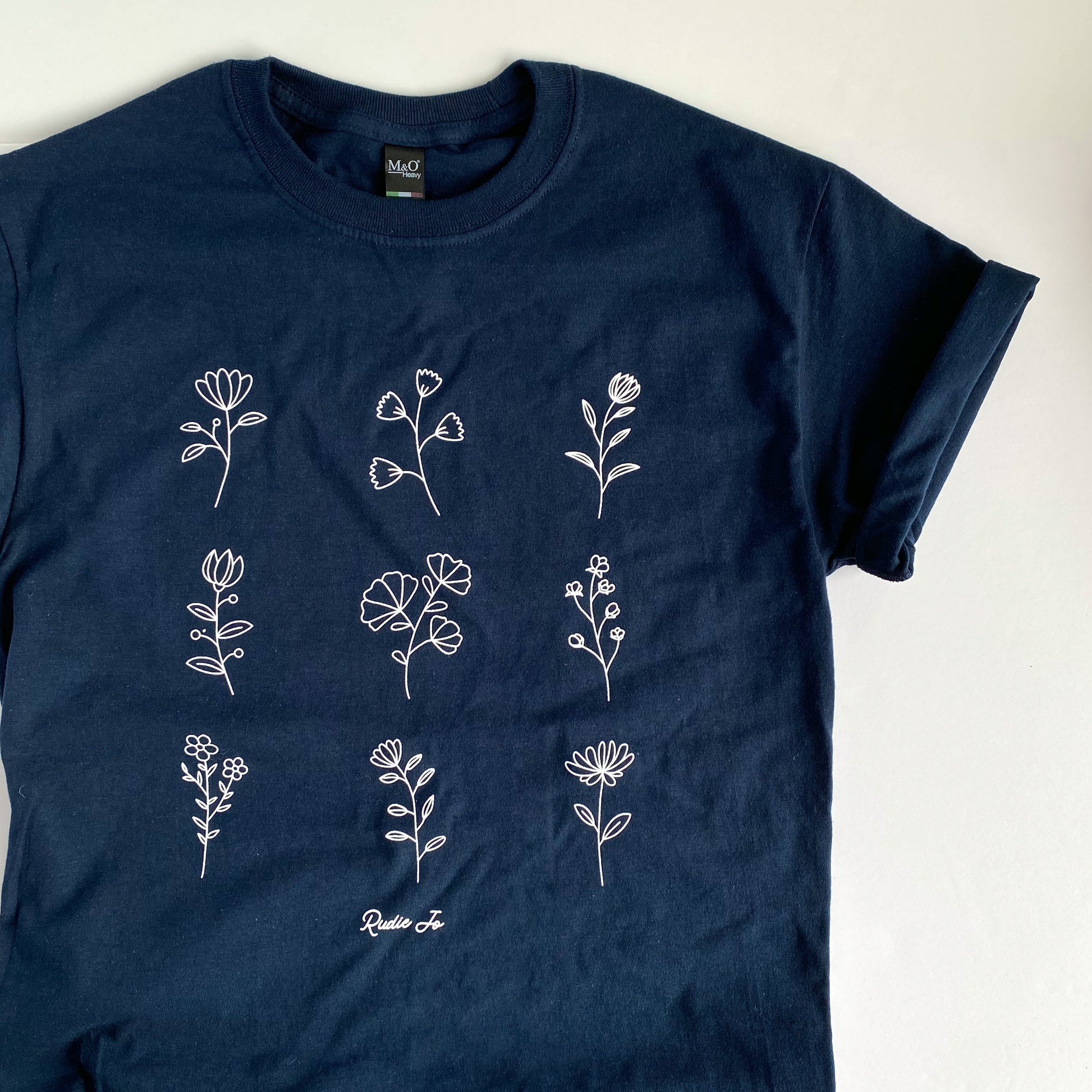 flat lay cotton navy blue t shirt with 9 white hand drawn floral stems printed on  the front in a 3x3 square with rudie jo logo below