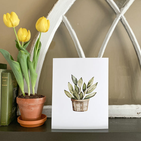 8x10 art print of green leafy stems in a brown basket on white background sits on a mantel next to a pot of 3 yellow tulips